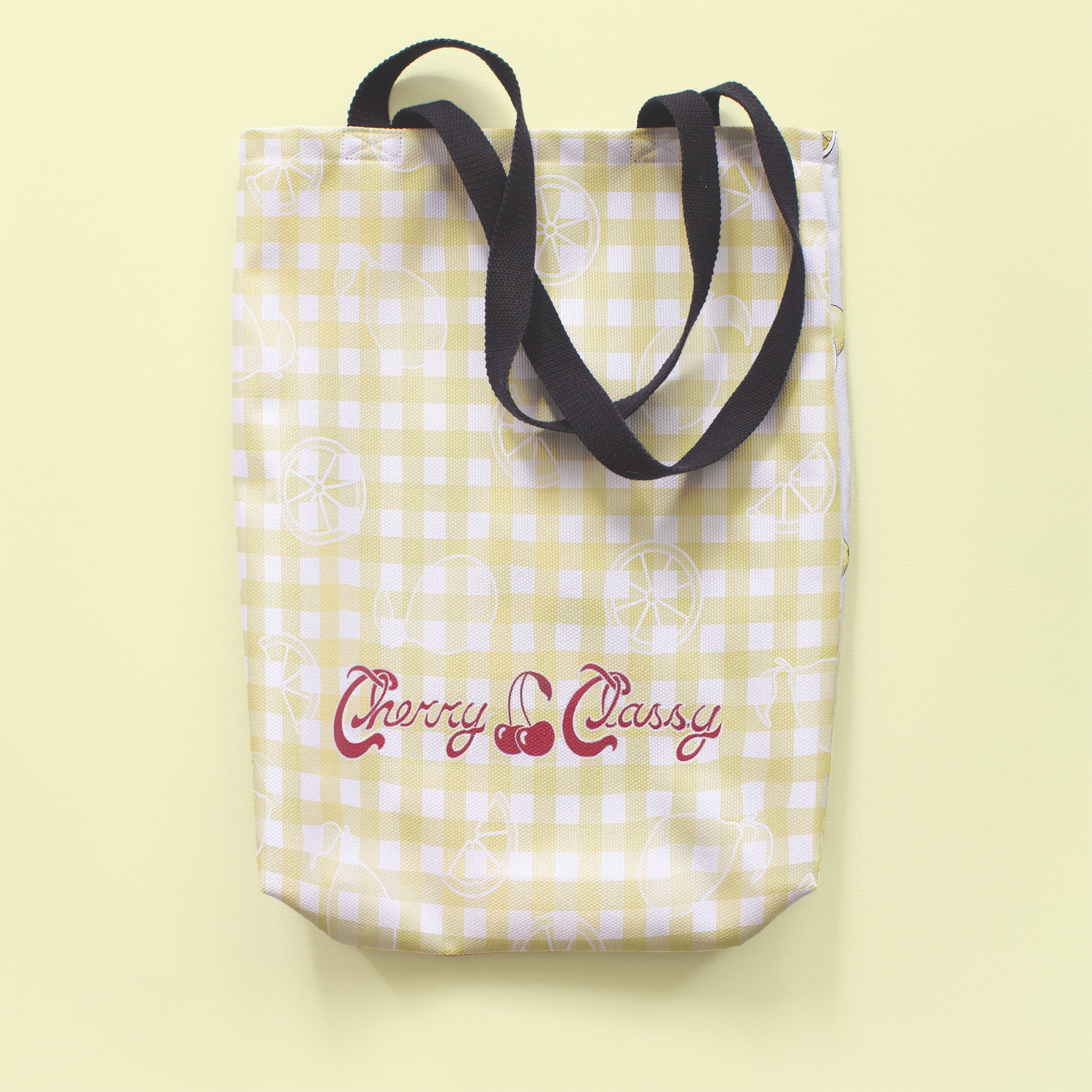 The back of the tote bag, featuring a yellow checkered pattern with a subtle white overlay of graphic lemons and vintage 'Cherry Classy' text on the bottom. The bag sits on a light lemon yellow background.